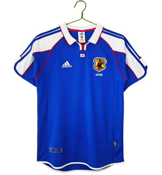 A blue and white short-sleeve soccer jersey with red trim, featuring the Adidas logo and the Japan national team crest, hanging on a green hanger against a white background.