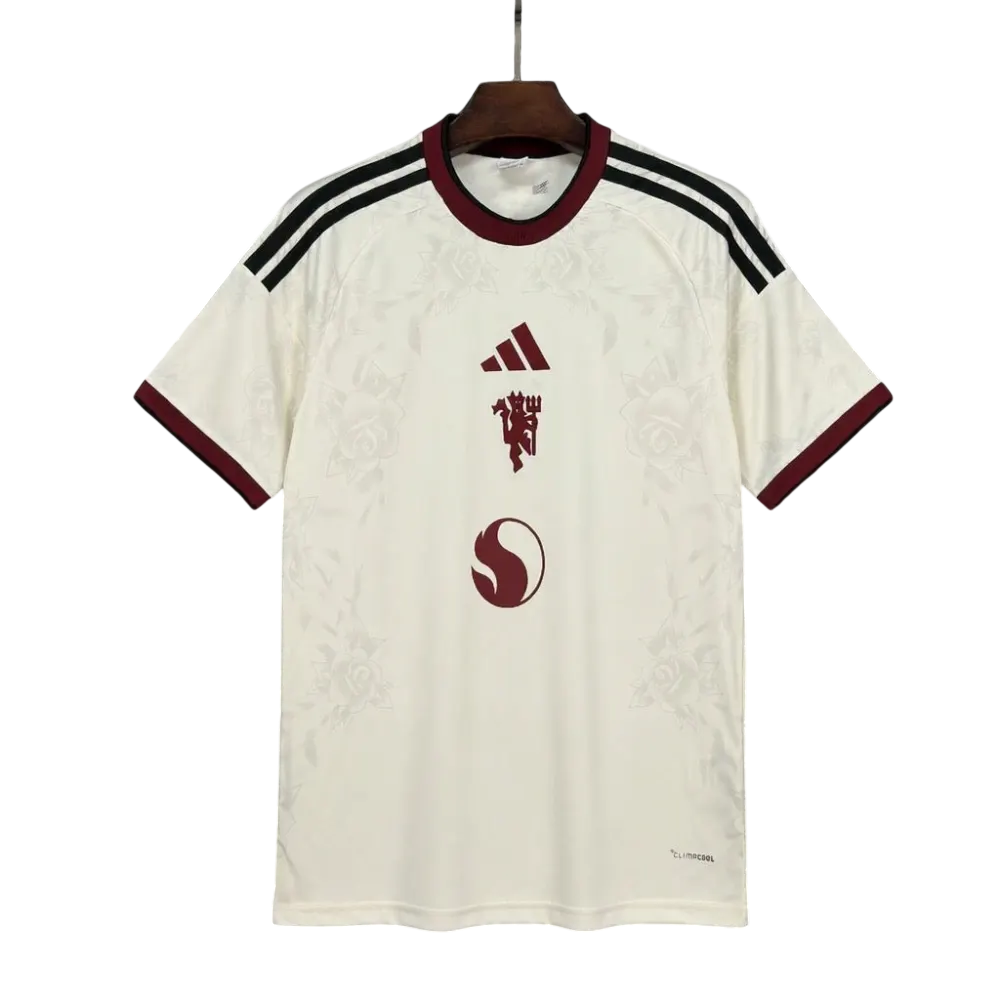 An Adidas Manchester United home soccer jersey in off-white, featuring a subtle rose pattern, dark red trim on the collar and cuffs, black and white shoulder stripes, and the club's crest with the Adidas logo and SFC crest on the front.