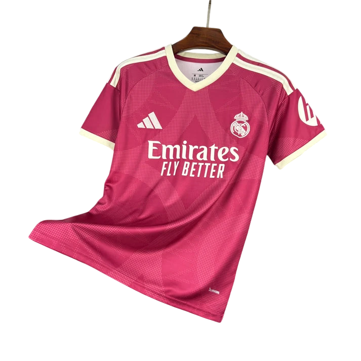 A Real Madrid home soccer jersey in a vibrant magenta-pink colorway, hanging on a wooden hanger against a plain white background. The jersey features cream-colored trim on the collar and sleeve cuffs, white Adidas branding, the Real Madrid club crest, and the Emirates Fly Better sponsor logo.